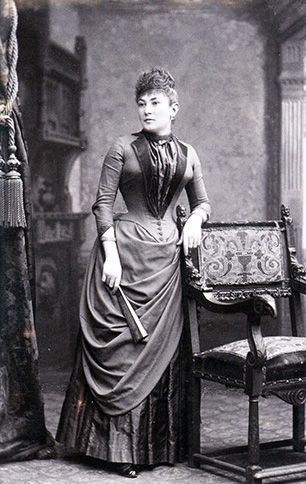 Portrait en pied d'une jeune femme en robe à tournure, un éventail à la main, appuyée sur une chaise sculptée devant un décor de studio